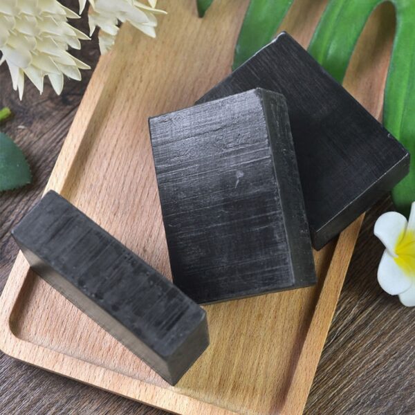 Three charcoal soap bars on a wooden plate with natural flower decor, showcasing handmade skincare aesthetics